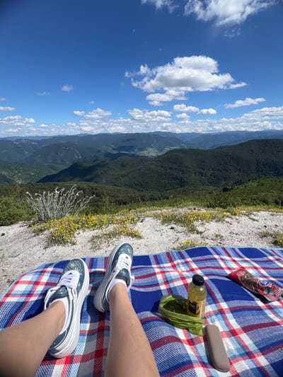 A pair of legs in white socks and blue and white sneakers rests on a blue and red plaid blanket spread on a rocky, grassy slope. Small yellow flowers dot the slope. Behind the blanket, a vast, green, mountainous landscape stretches to the horizon under a bright blue sky with scattered white clouds. A bottle of yellow liquid, a green bag, and a grey electronic device are on the blanket. A red and white package of snacks is also on the blanket. The location is identified as the city of Kosovo, Bulgaria.