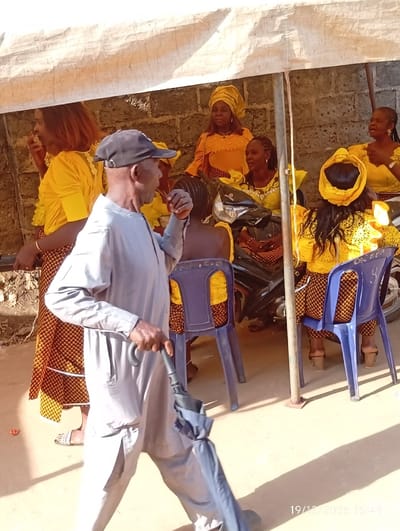 The image captures an outdoor social gathering in Isinyi-Nando, Nigeria, taking place under a temporary canopy made of light-colored fabric. In the foreground, an elderly male, wearing a grey long-sleeved tunic, matching trousers, and a dark baseball cap, walks from left to right, holding a partially folded dark umbrella downwards in his right hand. He appears to be looking towards the right side of the frame. Behind him, under the canopy, a group of approximately seven women are visible, most dressed in coordinated yellow attire, some with matching head wraps. Several women are seated on blue plastic chairs, while others stand or are partially obscured. A woman on the far left, wearing a yellow top and a patterned skirt, has her hand near her mouth. A black and silver motorcycle is parked behind some of the seated women. The background consists of a rough concrete block wall. The ground surface is light-colored, possibly concrete or packed earth, with shadows indicating overhead sunlight. A digital timestamp "19/12/2025 15:49" is visible in the lower right corner.