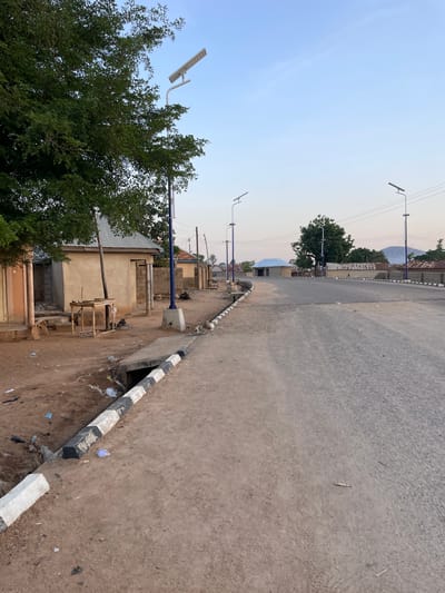 The image depicts a street scene in Bauchi, Nigeria. The road is paved and stretches into the distance, occupying the right side of the frame. A striped black and white curb runs along the edge of the road on the right. To the left of the curb is a dirt area littered with trash. Adjacent to the dirt area are buildings of varying construction, some with corrugated metal roofs. A large tree is visible on the left side of the frame. Streetlights with solar panels are spaced along the road, on the left side. Overhead power lines are also visible. In the distance, buildings and a hill are visible under a light sky. The scene is devoid of people.