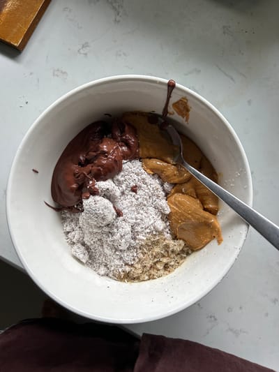 A white ceramic bowl sits on a white speckled countertop. Inside the bowl, there are several ingredients: a dollop of dark chocolate spread, a pile of shredded coconut, a mixture of what appears to be breadcrumbs or nuts, and two portions of peanut butter. A metal spoon is resting in one of the peanut butter portions. To the upper left of the bowl, a wooden object is visible. The overall scene suggests the preparation of food, possibly a recipe or snack. The location is Chang Phueak, Thailand.