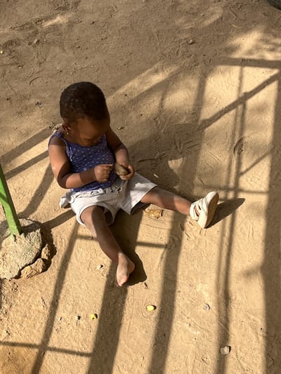 This high-angle outdoor photograph, taken in Zaria, Nigeria, shows a young child lying on a dusty, sandy ground under bright sunlight. The child, with dark skin and short, dark hair, is dressed in a blue or purple polka-dotted top and light-colored shorts. One foot wears a white sandal, while the other is bare. The child appears to be engaged in solitary play, holding a small, irregular, light-brown object (possibly a stone or seed pod) in their right hand, seemingly examining it with concentration.
The ground is textured with footprints and small indentations, scattered with small rocks, pebbles, and a few small green or yellowish objects that resemble seeds or nuts. A larger, light-colored rock is visible towards the bottom right, and a segment of a green metal bar or pipe runs along the bottom edge of the frame. Strong, linear shadows from an overhead structure, possibly a fence or lattice, are cast across the ground, indicating bright sunshine and suggesting either morning or afternoon. The scene conveys a sense of calm, simple outdoor play in an unpaved, possibly yard or open area. No text is visible in the image.