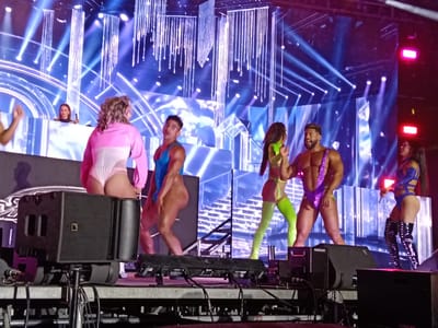 The image captures a stage performance, likely a concert or show, in San Bartolomé de Tirajana, Spain. Several performers are visible, dressed in revealing, colorful outfits. The backdrop is a complex arrangement of lights and digital displays. Stage equipment, including speakers marked "HURACAN," is prominent in the foreground. In the background, a DJ booth with a woman visible suggests a musical performance. The setting appears to be an outdoor venue or a large indoor space set up for a performance.