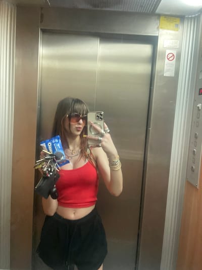 A person is taking a selfie in what appears to be an elevator. The elevator's interior is stainless steel. There is an indicator panel above, with buttons and floor indicators. Warning signage is visible on the upper section of the elevator door. The person taking the photo is wearing a red top, a black skirt, and sunglasses. They are holding an iPhone in their left hand and a pack of Oreo ice cream sandwiches with keys in their right hand. The person is wearing multiple rings and bracelets. There is no indication of the location of Petah Tikva, Israel, in the image.