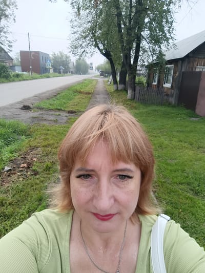 A middle-aged Caucasian woman with blonde hair and freckles is taking a selfie outdoors. She is wearing a light green ribbed shirt and a silver necklace. She has makeup on, including mascara and red lipstick. She is standing on a sidewalk next to a grassy area. In the background, there is a paved road with traffic signs and some buildings, including a wooden house with a fence. The environment appears to be a somewhat overcast day in a rural or semi-rural area, likely in Tugulym, Russia, given the architectural style of the buildings and the overall atmosphere.