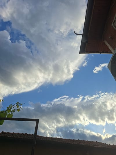An outdoor, upward-facing shot captures a bright, partly cloudy sky framed by sections of buildings and foliage in Galabovo, Bulgaria. The dominant feature is the expansive blue sky interspersed with large, fluffy white and grey clouds, some brilliantly illuminated by the sun, suggesting daytime.
In the upper left, the underside of a wooden eave or roof overhang is visible, featuring brown planks and a dark gutter from which a curved, dark pipe or hook descends. Part of an outdoor air conditioning unit, specifically its circular fan grille, is partially seen in the top left corner, set against the bright sky.
On the right side of the frame, a section of a building with textured, possibly terracotta, roof tiles is silhouetted against the sky, indicating it is largely in shadow. Below this, an L-shaped, dark metal bar or frame extends into the shot.
In the bottom right, a small cluster of vibrant green foliage, appearing to be from a vine with delicate tendrils and characteristic leaves, grows near the metal structure.
No people are visible. The scene captures a peaceful moment looking up from a likely residential setting, with bright daylight and a mix of clear and cloudy skies. There is no visible text in the image.