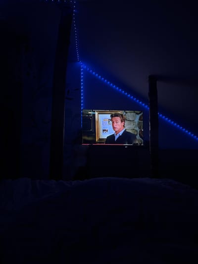 A dimly lit room with blue LED strip lights along the ceiling and wall. A television displays a man in a suit, possibly from a show or movie. A wooden beam is partially visible on the ceiling. The scene appears to be indoors, possibly a bedroom, with a bed visible on the right. No specific details indicate the location is Elhovo, Bulgaria.