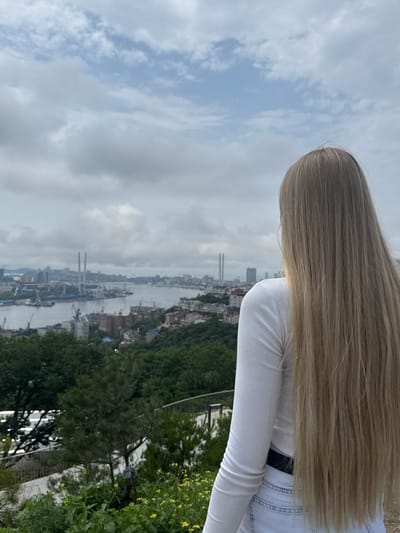 A blonde-haired individual wearing a white long-sleeved shirt and denim shorts stands facing away from the camera, looking out over the city of Vladivostok, Russia. The individual's hair is long and straight, reaching down their back. The city panorama shows a bay with a large bridge in the distance, as well as buildings, cranes, and trees. The sky is cloudy.