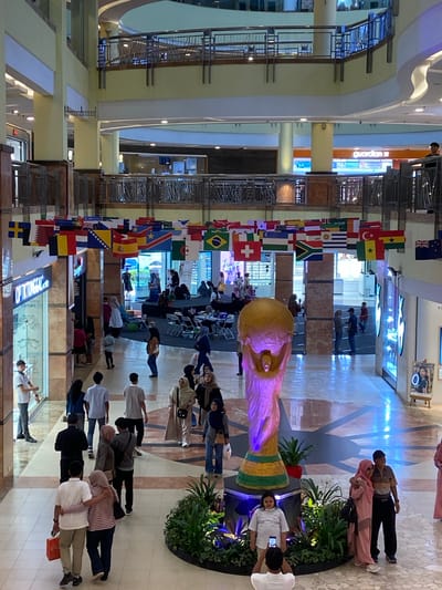 The image depicts an indoor scene, likely a shopping mall, with multiple levels visible. The central area features a large, golden sculpture, possibly a replica of a trophy, illuminated with purple and blue lights. The floor is tiled in a pattern of light brown and dark grey with radiating lines.
A prominent display of international flags hangs suspended across the upper levels, indicating a potential event or theme related to global sports or culture. Various people are scattered throughout the mall, engaged in activities such as walking, shopping, and socializing.
In the foreground, several individuals are seen walking or standing. To the left, a man in a white shirt and black pants is looking towards a store labeled "OPTICAL." Further down the walkway, a couple walks arm-in-arm, the man in a white shirt and beige pants, and the woman in a striped shirt and dark pants.
On the right side of the image, a couple is posing for a photo in front of the golden sculpture. The woman is wearing a pink hijab and dress, and the man is in a striped polo shirt and black pants. A young girl in a striped t-shirt stands to the left of the couple, facing the camera. Another person is taking a photo of the couple.
In the background, people are visible in various shops and on different levels of the mall. The architecture suggests a modern commercial building with metal railings and balconies on the upper floors. The overall atmosphere appears to be lively and public.
The presence of the "Optikal" store and the general mall layout suggests this location is likely a commercial center in Pekanbaru, Indonesia, which is known for its shopping malls. The specific mall is not identifiable from the image alone.