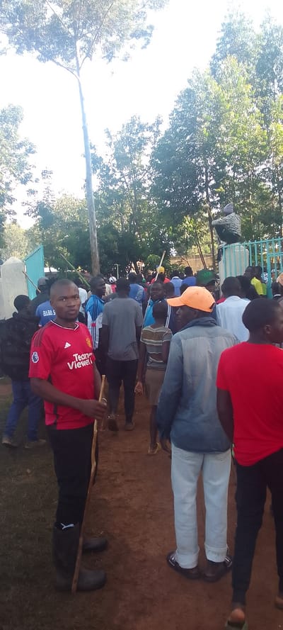 The scene is a gathering of people in a park-like area in Isukha ICHINA, Kenya (latitude: 0.351, longitude: 34.782). There is a large tree in the middle of the scene, with many trees surrounding it, and some people are standing on a dirt path. There are two people in the foreground, one is holding a stick and wearing a red shirt with the words "Team Viewer" printed on it, and the other is wearing a blue shirt and white pants. There are multiple people in the background, standing, sitting, and walking, with one person sitting on a fence. The people are mostly men and boys. There is a blue metal fence in the background. The ground is dirt with some grass, and the sky is bright blue with some white clouds. The atmosphere is casual and there are no signs of conflict. The people are likely gathered for a social event or gathering.