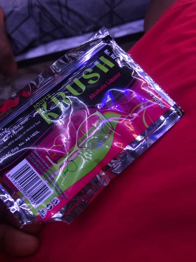 A close-up shot shows a foil packet for an alcoholic liqueur product, labeled "KRUSH," with a net volume of 55ml and 38% alc/vol. The packet is predominantly pink and green with silver accents and displays a barcode and "ADULTS ONLY 18+" icon. The packet is resting on a red fabric surface, possibly clothing. The background is out of focus, showing a person in dark attire and a wall with a textured surface and purple lighting. The image appears to be taken in a domestic setting, as there are no specific indicators of New Stadium, Nigeria.