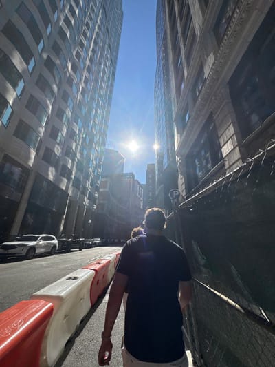 This image depicts a pedestrian-level view looking south down Congress Street in the Financial District of Boston, Massachusetts, United States. The clear blue sky shows a prominent sunburst with lens flare, indicating a bright, sunny day.
On the left side of the street, the modern, multi-story skyscraper at 100 Federal Street rises, characterized by its distinctive curved, reflective glass facade and numerous windows. Adjacent to the street, a line of interconnected red and white plastic construction barriers separates the roadway from parked vehicles. Several cars are visible, including a white SUV parked closest to the foreground.
On the right side, an older, tall building with a dark, ornate stone or brick facade features classical architectural details, including cornices and multiple rectangular windows. A dark mesh-covered chain-link fence runs along the sidewalk in front of this building, likely indicating a construction or renovation site. A blue circular "P" parking sign is affixed to a pole or the fence.
In the foreground, a male individual is walking away from the camera. He has short, curly brown hair, wears a dark short-sleeved polo shirt, and light-colored shorts or pants. His left arm is visible, showing a bracelet or watch. A second person is partially obscured behind his left shoulder. The street is asphalt, and visible shadows are sharp due to the direct sunlight.
