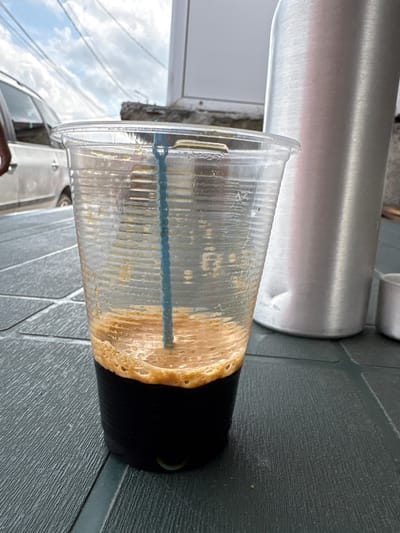 The image captures a partially consumed beverage in a clear plastic cup, resting on a dark grey, textured surface. The cup contains a small amount of frothy, light brown liquid, likely coffee or a similar iced drink, with residue clinging to the inner walls above the liquid line. A blue, ridged straw is visible in the drink. The bottom portion of the plastic cup is an opaque black.
In the background, a large, cylindrical object made of brushed silver metal, possibly aluminum, extends horizontally behind the cup. To the left, reflections of a cloudy sky and what appear to be power lines or wires are visible, suggesting an outdoor setting. A light-colored vehicle, possibly a car, is partially visible further in the background. The scene depicts the aftermath of someone enjoying a drink on a daytime outing. There is no discernible text visible in the image. The setting appears to be a casual outdoor area in Karadjovo, Bulgaria.