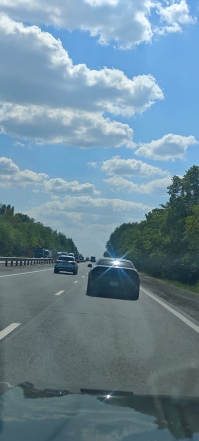 The image shows a view from inside a car on a highway, looking forward. Other cars are visible ahead, with the closest car being a dark-colored sports car. The highway is bordered by trees on both sides. Above, the sky is blue with numerous white clouds. The city of Bely Klyuch, Russia, is not identifiable from the image.