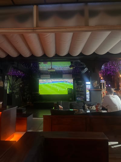 The image shows the interior of a dimly lit establishment, likely a bar or restaurant, in Thủ Đức, Vietnam. A large projection screen displays a soccer match, with green turf and players visible. The ceiling features white, cushioned, accordion-style coverings. Ambient purple lighting is visible in the background, along with strings of small purple lights. To the left, a sign reads "SKY 9". A person wearing a white shirt is seated facing away from the camera, looking towards the screen. Various seating areas, tables, and what appear to be drink dispensers or refrigerators are present in the environment. The scene suggests a casual atmosphere for watching sports.