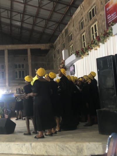 The image depicts a group of people, seemingly a choir, wearing black robes and yellow hats. They appear to be on a stage or elevated platform. The background indicates an indoor space with a ceiling featuring rows of recessed lights, possibly a hall or church in Nteje, Nigeria. There is floral decoration above the stage. Some speakers are visible near the front of the stage.