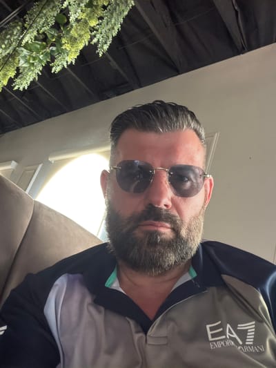 A person wearing sunglasses and an Emporio Armani EA7 athletic jacket is sitting on a brown couch. The location appears to be indoors, possibly a residence in Spring Valley, United States of America, though specific evidence supporting this location is not visible in the image. The person is facing the camera with a slight angle. A plant is visible in the background, near a white pillar. Artificial lighting is present.