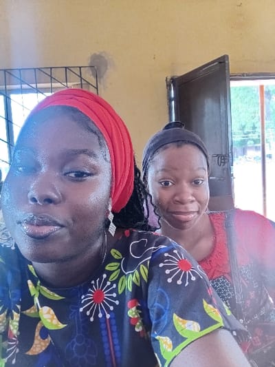 The image depicts two adult females positioned indoors in the city of Ejule, Nigeria. The foreground individual, dark-skinned, wears a red head covering, a multi-colored patterned shirt, and a silver-toned dangling earring on her visible left ear. Her lips appear glossy, and her gaze is directed towards the camera. Behind her and to the right, a second dark-skinned female is partially visible, wearing a dark headband and a reddish top, presenting a slight smile. The indoor background consists of light yellow-painted walls, one showing a grey plaster repair patch. To the left, a window with a dark metal security grille is visible, allowing light into the room. To the right, a dark brown door stands ajar, revealing an outdoor scene with green foliage and ambient light beyond.