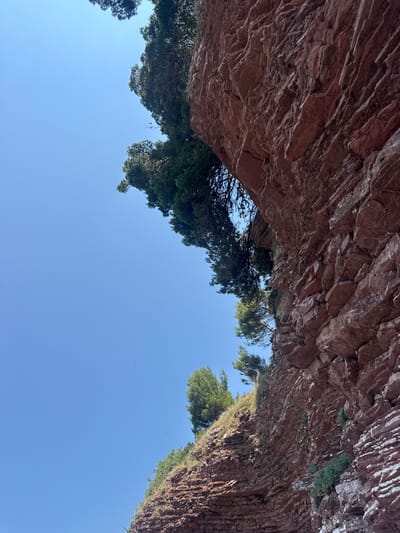 The image depicts a low-angle view looking upwards towards a rocky overhang and a clear blue sky. The rock formation is reddish-brown with visible stratification and some green vegetation growing in crevices. Lush green trees with dense foliage are seen peeking out from behind the rock, some appearing to be pine trees. The overall scene suggests an outdoor natural environment, possibly a coastal area or a canyon, given the rocky terrain and the presence of trees. The bright blue sky indicates a sunny day, likely midday or afternoon. There are no people or specific objects visible, focusing solely on the natural elements of the landscape. The composition creates a sense of being sheltered beneath the rock, looking out into the open sky. The location context provided is Pržno, Montenegro, which aligns with a Mediterranean-like landscape with rocky coastlines and vegetation. There is no visible text in the image.