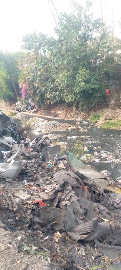 The scene depicts severe environmental degradation along a waterway in Thika Township ward, Kenya. In the foreground, a substantial mound of refuse is accumulated, primarily composed of fragmented automotive parts, including plastic bumper sections, vehicle interior panels, and metal components, interspersed with general debris. Adjacent to this, a dark, murky body of water, consistent with a river or stream, flows, heavily laden with floating garbage such as plastic bottles, bags, and various indeterminate synthetic and organic waste. The banks of this waterway are visibly eroded and also covered in concentrated refuse. The background features a dense line of green trees and bushes on a raised bank, with some pink-purple flowering plants visible. A red cylindrical object is discernible within the vegetation on the distant bank. No individuals are present in the image, and no active interactions or actions are depicted.