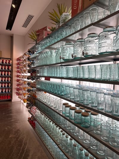 The image is a low-angle shot looking up at a ceiling display of Coca-Cola-themed glass bottles and merchandise in the Coca-Cola Store, located in Lake Buena Vista, Florida, United States. The bottles are arranged in neat rows on metal shelves that run along the ceiling, giving the impression of a grid or a gallery of glass. Some of the bottles are clear, bearing the Coca-Cola logo, while others appear to be mason jar-style glasses with the Coca-Cola logo and spouts. Further down, rows of Coca-Cola merchandise, including mugs and possibly small decorative items, are suspended from the shelves. At the far end of the image, a wall display of Coca-Cola-branded products is visible, appearing to be footwear or clothing. The environment is well-lit, and the perspective emphasizes the symmetry and repetition of the items on display.