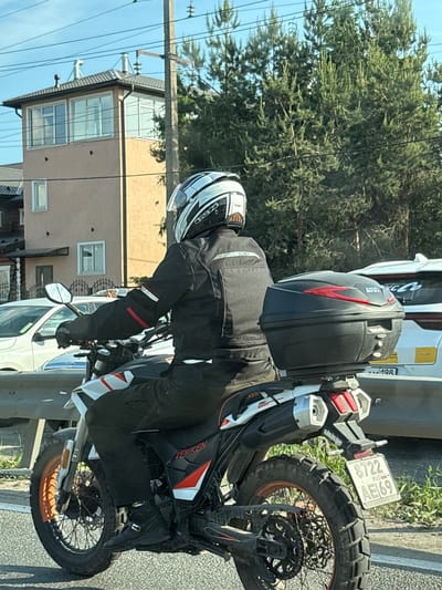 A motorcyclist is observed from behind, stationary or moving slowly on a paved road, under clear skies. The scene is set in Novosaratovka, Russia. The motorcyclist wears a white, black, and grey full-face helmet with a clear visor (brand "SOLEH" visible on the rear), a black motorcycle jacket with white and red accents (logo "MCJ" on the back), black pants, and black buckle-fastened motorcycle boots.
The individual is operating a black and white "TEKKEN" brand adventure-style motorcycle with red/orange accents. The front wheel features an orange rim and knobby tire, while the rear wheel has a black rim and a similarly knobby tire. A black "GIVI" brand top case with red accents is mounted on the rear luggage rack. The motorcycle's rear fender displays a Russian license plate: "8722 RUS AE69", with "69" indicating the Tver Oblast region.
In the background, to the left, a light brown three-story building with a dark grey hip roof is prominent. The top floor features large, white-framed windows suggesting an observation area, and two metallic ventilation pipes and a chimney are visible on the roof. Lower down, additional white-framed windows are visible. A white car, possibly a sedan, is partially visible to the left of the motorcycle, behind a metal guardrail. To the right, dense green coniferous trees are present, partially obscured by a tall wooden utility pole supporting multiple power lines. Further back, another white vehicle, likely an SUV, has a "MC Co" sticker on its rear window and "198" faintly visible on its bumper. The road surface is asphalt.