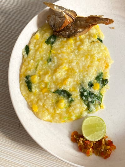 An overhead shot displays a plate of a ready-to-eat meal, likely a local Indonesian dish, against a striped placemat. The main component is a creamy, yellowish porridge, interspersed with bright yellow corn kernels and dark green leafy vegetables, resembling spinach.

To the left of the porridge are two small pieces of fried fish, golden-brown and crispy in appearance. On the right side, a fresh lime wedge and a small serving of vibrant red chili relish, or sambal, complete the presentation. The plate itself is off-white with a subtle textured finish.

The setting appears to be a casual dining environment, possibly at home. The arrangement suggests a meal prepared for consumption. The specific combination of ingredients—a savory corn and vegetable porridge served with fried fish, lime, and sambal—is characteristic of Indonesian cuisine, particularly dishes like Tinutuan (Manado Porridge) often found in regions such as Bekasi Regency, Indonesia. There is no visible text in the image, and the time of day or specific emotions are not discernible.