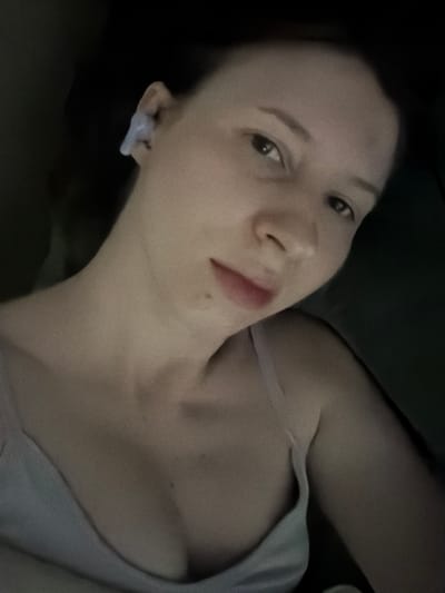 A woman is lying down, looking into the camera. She has on a light-colored tank top and is wearing an earbud in her left ear. The lighting is dim, with the majority of the image being dark, but the woman's face and upper body are illuminated. The background is dark and out of focus, making it impossible to determine the location.