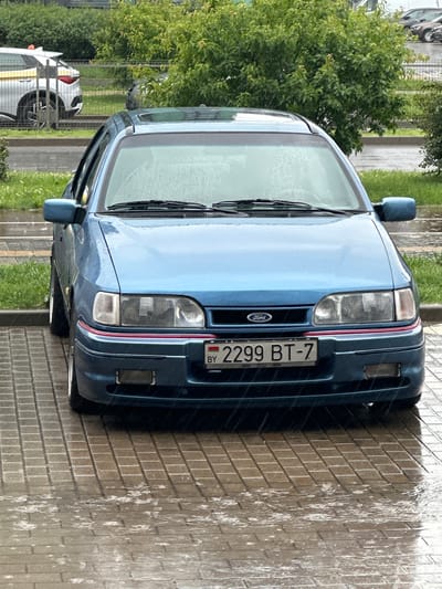 The image captures a blue Ford Sierra parked on a wet, tiled surface in what appears to be an outdoor setting. Rain is visible falling, indicated by streaks on the car and puddles on the ground. The car is the primary subject, with its front prominently displayed. The license plate reads "2299 BT-7" with "BY" in the corner, suggesting it is registered in Belarus. In the background, there are green trees and bushes, as well as a glimpse of other vehicles and a fence. The overall impression is of a car parked in a city during a rain shower.