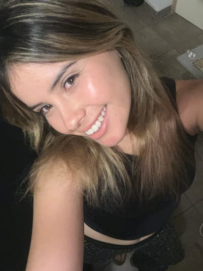 A selfie photograph showing a woman with long, light brown hair smiling with white teeth. She is wearing a black top. The location of the image is not specified, and there is no reference to Sumalao, Argentina, in the picture. The background is indistinct but appears to include dark-colored clothing and possibly a floor.