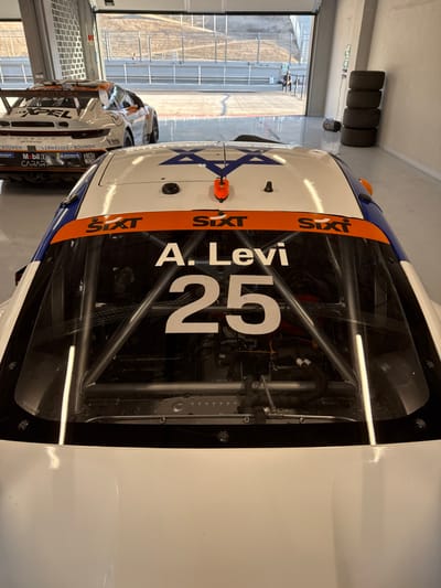 A white race car with a blue and orange stripe is angled. The car displays "A. Levi" and the number "25" in white lettering on the side window. The Star of David is on the hood. "Sixt" is visible in orange on the side. Additional race cars are visible in the background. A set of tires is stacked against the wall to the left of the vehicle.