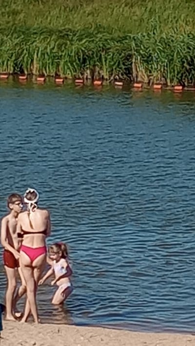 The image shows a group of people sunbathing by a body of water. There are at least three individuals visible, two of whom are lying down and one who appears to be sitting or lying partially in the water. One person is wearing a red bikini and another is wearing a white top and dark bottoms. The setting is a grassy area next to a blue, rippling body of water, possibly a lake or pond, with a sandy or dirt bank on the right side. The sun is shining brightly, suggesting it is daytime and likely warm weather, ideal for sunbathing. The people are relaxed, enjoying the sun. The image is taken from an overhead perspective, looking down at the scene. There is no visible text in the image.