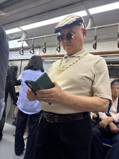 The image shows several people on a subway car in Seoul, South Korea. The main subject is an elderly man wearing a light-colored polo shirt, black pants, a brown belt, and a striped cap. He is also wearing sunglasses and a necklace made of light-colored beads. He is holding a dark green phone or wallet in his right hand, and a ring with an emblem is visible on his left hand.
To his left, a younger person with dark hair is wearing a blue and white striped shirt. Further in the background, another person is asleep, leaning their head on their hand. The subway car has metal poles with brown handles for passengers to hold onto, and bright fluorescent lights illuminate the interior. The overall scene suggests a typical commute during the day.
No visible text is clearly decipherable, though the belt buckle appears to have some markings.