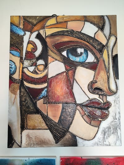 The image shows a square painting of a face in a cubist style. The face is depicted in various geometric shapes and colors, with outlines in what appears to be gold or copper. The eye is a prominent blue, and other colors include browns, oranges, whites, and blacks. The painting is hanging on a white wall. In the lower part of the photo, parts of two other paintings can be seen.
There is no information in the image to indicate that it is located in Seixal, Portugal.