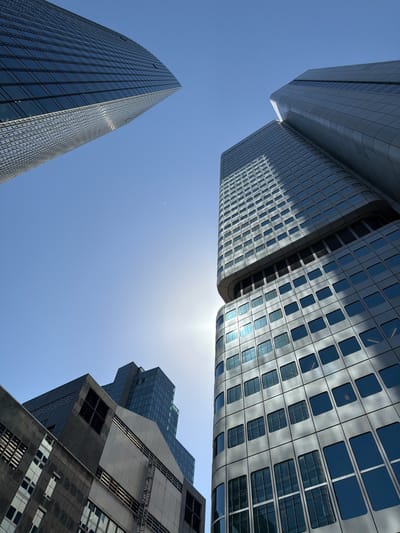 This media file captures a low-angle view of several modern skyscrapers against a clear blue sky. The architecture is characterized by sleek glass and metal facades, with multiple buildings reflecting the sunlight. One building on the left is a tall, triangular skyscraper with a grid-like pattern of windows. In the center and right, two other buildings are visible. One has a curved, metallic exterior with rows of square and rectangular windows, showing reflections of the sky and other buildings. Another building on the far right features a more complex, angular design with dark, reflective surfaces and distinct sections of windows. The sun is visible, high in the sky, indicating it is likely midday. The overall impression is one of urban grandeur and contemporary architectural design, with the clear sky suggesting fair weather. There is no visible text in the image. The scene is set in Frankfurt, Germany, known for its prominent skyline dominated by high-rise buildings.