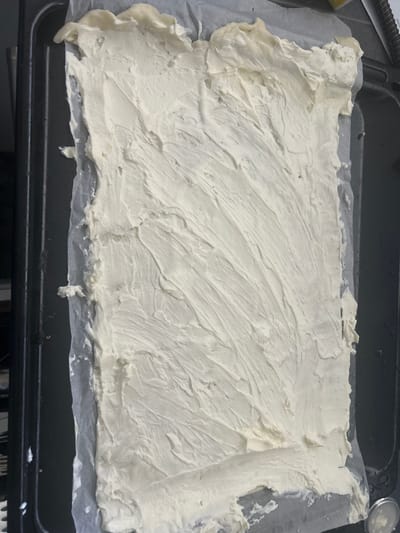 An overhead, close-up shot captures a baking sheet lined with parchment paper, upon which a thick, off-white frosting or cream cheese mixture has been spread unevenly. The mixture is textured with visible strokes from a spreading utensil, suggesting it is either a base for a dessert or a filling. The baking sheet itself is dark gray and appears to be made of metal. The scene is indoors, with a dark, indistinct background suggesting a kitchen or food preparation area. No specific landmarks or contextual elements point to the city of Leiden, Netherlands. The image is devoid of any human presence or interaction.