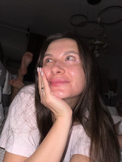 A young person with long, dark brown hair and light-colored eyes is depicted in a casual, indoor setting, likely Akulovo (Gp Kubinka), Russia. They are lying down, wearing a light-colored t-shirt, and posing for the camera with their right hand resting under their chin. A ring with multiple bands is visible on their right ring finger, and their fingernails are painted with a light polish. Their skin appears somewhat shiny with visible blemishes and redness. In the background, their bare feet are visible, elevated. The lighting is dim, suggesting it is evening or night. Blurry shapes in the background might be decorative elements or light fixtures. The person has a relaxed, possibly playful expression.