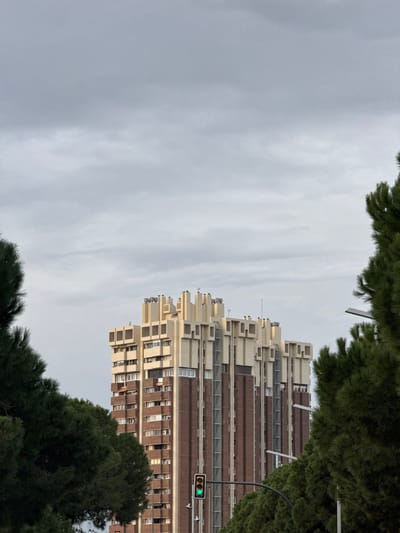 A tall building, featuring a unique staggered design with beige and brown color tones, dominates the view. It's partially framed by green trees at the top and bottom. A street signal with a lit green light is visible near the building. The background is a cloudy sky. The perspective is from below, looking upwards at a slight angle.