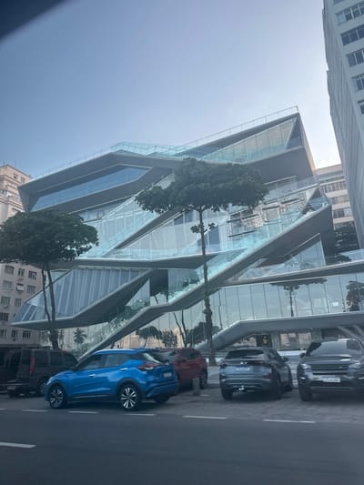 The original image, taken from a slightly low-angle perspective and rotated 90 degrees clockwise, displays an urban street scene in Rio de Janeiro, Brazil, on a clear, sunny day.
The dominant subject is a striking, modern multi-story building characterized by a complex, geometric facade of dark grey or black panels interspersed with large, reflective glass sections. These glass segments form diagonal and angular patterns, creating a dynamic and contemporary aesthetic. Portions of the glass reflect the blue sky and trees, while others reveal interior structures or architectural elements.
In the foreground, a city street runs parallel to the building, featuring several parked cars. Most prominently, a bright blue SUV is parked closest to the viewer. Behind it, a dark grey or black SUV, a black SUV, and a dark red car are also visible. Green-leafed trees are scattered along the street and beside the building, adding natural elements to the concrete and glass environment.
In the background and to the sides, older, more traditional buildings with numerous windows can be seen, contrasting with the modern architecture of the main structure. The sky is a clear, light blue, indicating fair weather and daytime. No people are explicitly visible, and no discernible text apart from illegible license plates on the vehicles. The scene captures a vibrant blend of contemporary and established urban landscapes.