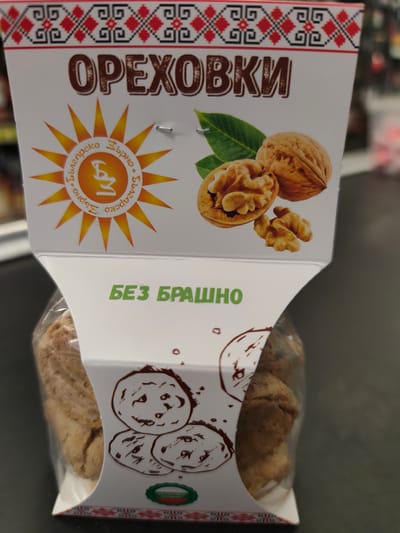 The image shows a package of baked goods labeled "ОРЕХОВКИ" (orekhvki, meaning "walnut cookies") and "БЕЗ БРАШНО" (bez brashno, meaning "flourless"). The packaging features a traditional Bulgarian folk pattern along the top and bottom edges. In the center, there is a stylized sun logo with Cyrillic text that appears to say "Българско зърно" (Balgarsko zarnyo, meaning "Bulgarian grain"). Images of walnuts and leaves are also present. Below the main label, the package contains several round, cookie-like items wrapped in clear plastic. A small circular emblem at the bottom of the package shows the Bulgarian flag and the text "НАТУРАЛЕН ПРОДУКТ" (naturalen produkt, meaning "natural product"). The product is displayed on a dark surface, likely a shelf in a retail environment. The image does not contain any identifiable landmarks or contextual clues to suggest its location within the city of Kazanlak, Bulgaria.