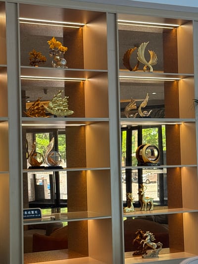 The image displays a well-lit shelving unit filled with various decorative items. The shelving unit appears to be located in a commercial establishment or a showroom, possibly in Linghe, China, given the presence of Chinese text on a sign.
On the top shelf, there are two golden tree-like ornaments, one smaller and to the left, and a larger one with fruit-like embellishments to the right. Next to the larger tree ornament is a golden elephant figurine.
The second shelf from the top features two abstract golden sculptures on the left, appearing to be wing-like or flame-like forms. On the right side of this shelf, there are two more abstract golden sculptures with spherical elements at their base.
The third shelf down showcases a golden sailing ship model on the left. On the right side, there is a circular golden decorative piece with concentric rings. Below this, a sign with Chinese characters is visible, likely indicating a service or designation.
The fourth shelf from the top displays two golden deer figurines on the left, one standing and one lying down. On the right side of this shelf, there are two golden horse figurines, one more prominent and detailed than the other, appearing to be in motion.
The environment appears to be indoors, with ambient lighting from fixtures within the shelving unit, casting a warm glow on the objects. The background, visible through the reflections on the shelves, suggests a window or glass partition showing greenery and possibly an outdoor scene with vehicles, consistent with an urban setting in Linghe, China. The shelving unit itself is constructed with light-colored wood or laminate, with glass shelves in some sections.