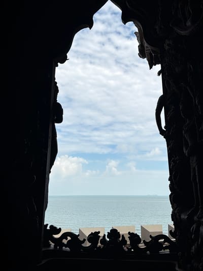 This image captures a view of the sea and sky through a intricately carved wooden archway. The archway is dark and silhouetted, obscuring much of the detail of its carvings, which appear to depict figures and ornate patterns. Through the opening, a calm sea with gentle waves stretches to the horizon, where a faint outline of land or a ship can be seen. The sky above is a mix of pale blue and white clouds, with the light suggesting it is daytime. In the foreground, a decorative wooden railing with a swirling, organic design runs along the bottom of the archway. The overall impression is one of peaceful contemplation of nature framed by artistic craftsmanship. This scene is characteristic of the Sanctuary of Truth in Pattaya City, Thailand, a magnificent wooden structure adorned with intricate carvings that overlooks the sea.