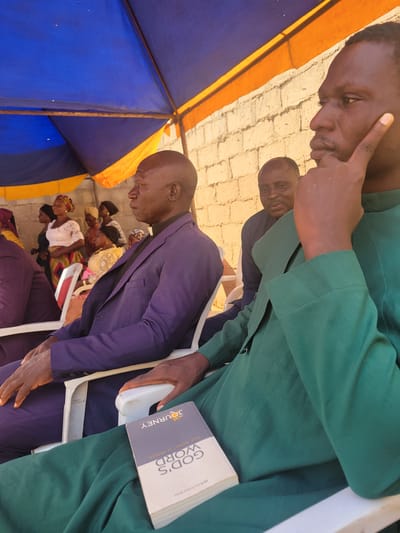 In Madalla, Nigeria, under a blue and orange tent, a group of people are gathered. A man in a purple suit and a white clerical collar sits in the foreground, facing left. To his right, another man in a green robe is seated, holding a book titled "GOD'S WORD YOUR JOURNEY TO FREEDOM." In the background, other men and women are visible, some standing and others seated. The setting appears to be outdoors, with a brick wall behind the tent. The overall atmosphere suggests a religious gathering or service.