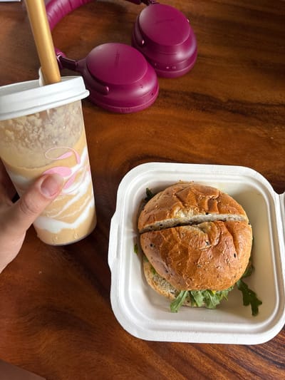 A top-down, eye-level, medium shot captures a meal on a wooden table, likely in the city of Ban Kanok 23, Thailand. In the foreground, a hand with short, unpolished fingernails grips a clear plastic cup filled with a layered beverage, possibly a smoothie or iced coffee, with a reusable golden straw inserted. The cup has a white lid and a pink swirled design. To the right of the drink, a white compostable clamshell container holds a halved sandwich on a sesame seed bun, revealing green arugula and what appears to be a light-colored spread. Behind the food and drink, a pair of magenta Bose wireless headphones rests on the table. The wooden surface exhibits a visible grain pattern.