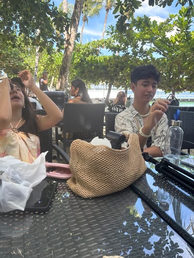 In Samal, Philippines, a young man and a young woman are seated at a table outdoors, likely at a restaurant with a view of the ocean. The man, with dark hair, is wearing a white shirt with a black line pattern and a beaded necklace and bracelet. He holds a small, light-colored object in his right hand. Next to him, a clear water bottle with a blue cap is visible on the table.
The woman, with long dark hair, is wearing a patterned halter top. Her mouth is open, and her hands are raised as if she is speaking or reacting to something. In front of her is a white paper bag and a pink pouch. A large, woven straw bag sits on the table between them, obscuring some of the items. The table appears to be made of dark, textured material, possibly glass with a woven pattern. In the background, palm trees and lush green foliage suggest a tropical environment. The ocean and a clear blue sky with some clouds are visible beyond the trees. Other people are seated at tables in the background.