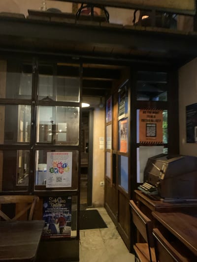 The image displays the dimly lit interior of an establishment, characterized by a prominent wall of glass-paned display cases or windows. No people are visibly present.
On the right side of the frame, dark wooden shelves or furniture are seen, with a vintage-style typewriter resting on one of the upper shelves. The floor appears to be a light-colored tile, with a dark mat visible in the background. The scene's lighting suggests it is either evening or an indoor area with restricted natural light, with reflections of internal light sources visible on the glass.
Several posters and signs are affixed to or reflected in the glass surfaces. An orange sign on the upper left, when correctly oriented, reads "DO YOU WANT FREE TATTOO QR CODE" and features a QR code. A white poster displayed on the central-right glass panels shows various social media logos (Facebook, Instagram, YouTube, TikTok, Pinterest, Twitter) with the Greek text "Συνδέσου μαζί μας" (Connect with us) and another QR code. To the far right, a blue poster advertises "Santa Claus Kingdom" with additional Greek text stating "ΤΟ ΠΙΟ ΠΑΡΑΜΥΘΕΝΙΟ ΧΡΙΣΤΟΥΓΕΝΝΙΑΤΙΚΟ EVENT ΤΗΣ ΑΘΗΝΑΣ!" (THE MOST FAIRYTALE CHRISTMAS EVENT OF ATHENS!). The scene is static, with no discernible activity taking place.