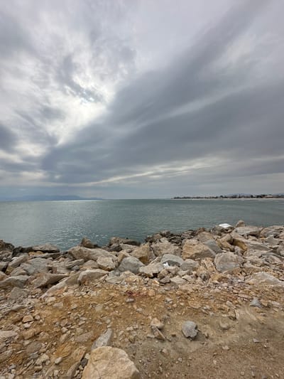 The scene depicts a coastal landscape in Limenas Avdiron, Greece, under an overcast sky. The foreground consists of rough, reddish-brown soil and gravel, transitioning into a rocky shoreline composed of numerous irregularly shaped boulders and stones in shades of gray, beige, and white. A small, dark reddish stain, approximately 15cm in diameter, is visible on the soil in the lower-middle-left quadrant. A crumpled white object, possibly paper or plastic, rests among the rocks on the right side of the embankment. The mid-ground features a calm body of water, likely the sea or a bay, reflecting diffused light from the sky. In the far background, a hazy landmass or mountains are visible on the left horizon, while a distant coastline with scattered low-rise structures extends along the right horizon. No individuals or active subjects are present.