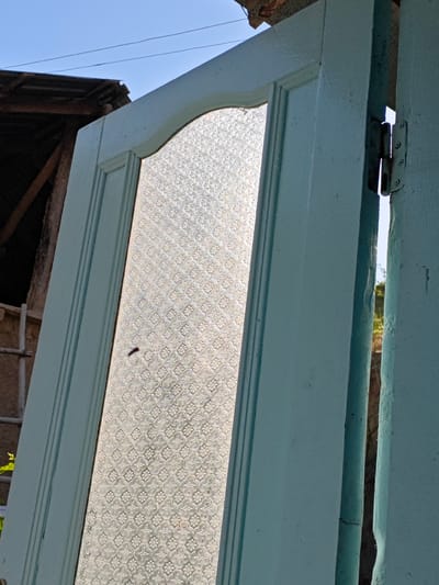 An outdoor, close-up, low-angle shot captures a portion of a light blue, wooden door frame and a section of the door itself, which is hinged to the frame. The door contains a large pane of decorative, textured glass that is illuminated by sunlight, creating a luminous effect. The sunlight also causes a shadow of a small insect, likely a bee or a fly, to be visible on the glass. The background is blurred but shows elements of an outdoor environment, including a section of a rustic roof with corrugated metal and wooden beams, as well as a clear blue sky. The scene is set in Chorbogh, Tajikistan.
