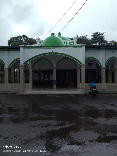 The image shows the front facade of a mosque, located in Kecamatan Haurgeulis, Indonesia. It is a modern structure with a white exterior and a green dome. The roof is covered in a green and white pattern. The facade has a series of arches supported by pillars, creating a covered walkway leading to the entrance of the mosque. There is a blue mailbox on the right side of the entrance. There is a motorcycle parked on the right side of the image. The ground is wet from recent rain, with puddles reflecting the sky. The sky is overcast with grey clouds.