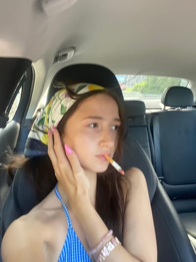 The image shows a young woman sitting in the passenger seat of a car. She is wearing a blue and white striped top and a patterned headscarf. She has bright pink fingernails. A lit cigarette is held between her lips. Her right hand is raised to her face, with her fingers touching her cheek. The interior of the car is visible, including the black leather seats and the headliner. Outside the car, trees and buildings can be seen through the windows, suggesting an urban or suburban environment. The lighting indicates it is daytime.