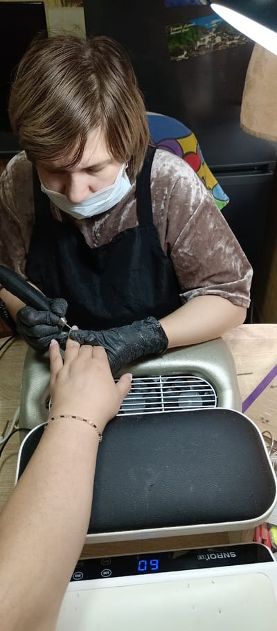 A manicurist wearing a light-colored, velvet-like shirt, a black apron, and a disposable face mask works on a client's nails, using an electric file. The manicurist wears black gloves. The client's arm is extended towards the manicurist, and she wears a beaded bracelet. The client's hand rests on a silver-colored nail dust collector with a grill. Below the collector, a white nail curing lamp with a black padded top displays a timer reading "09." The setting appears to be a room, with a black surface visible behind the manicurist, possibly a refrigerator or cabinet, with a colorful poster or decorative item attached. The location appears to be Irbit, Russia, based on the poster.